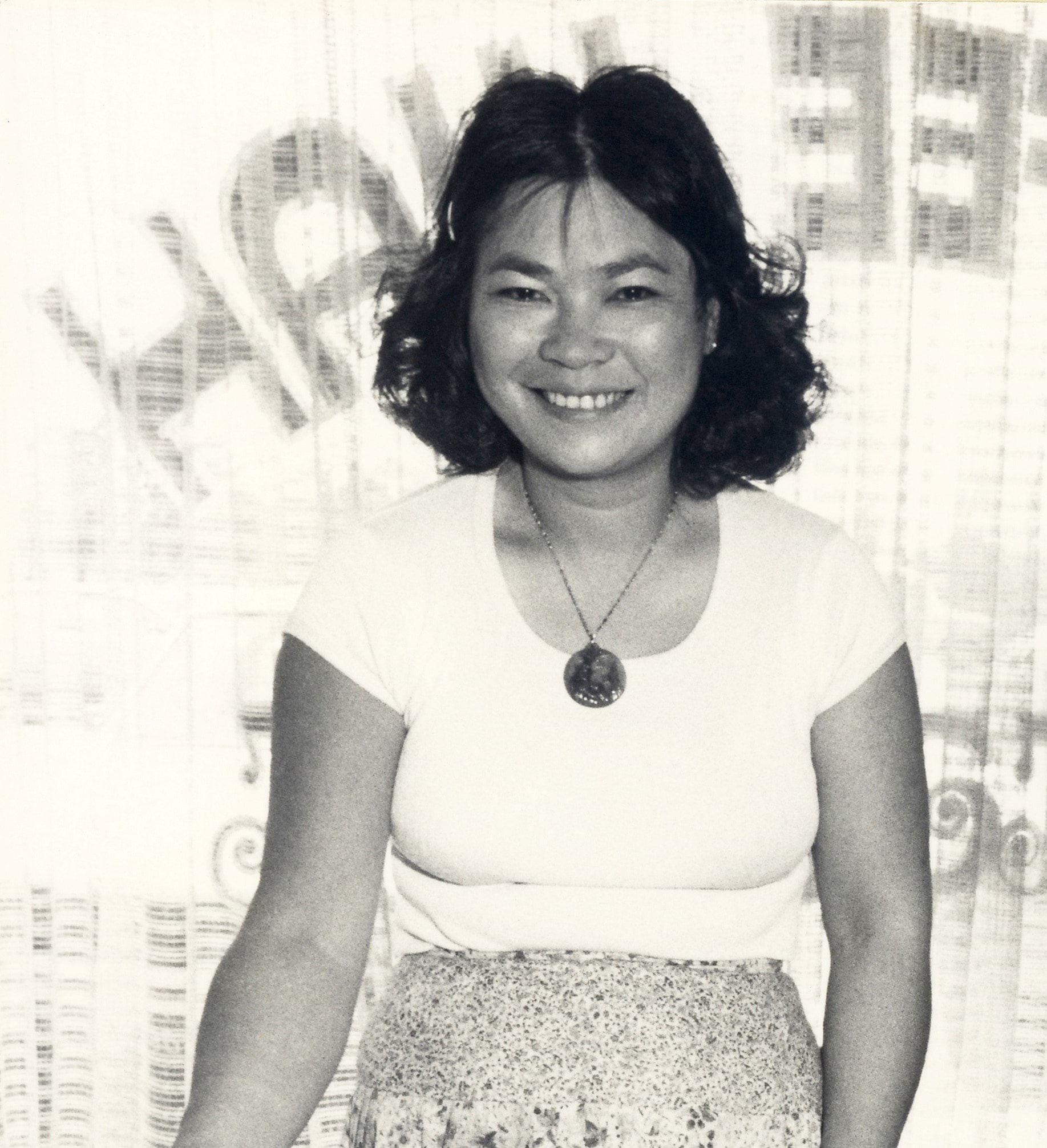 Black and white photo of Jenny Rose Ng smiling at the camera, wearing a white t-shirt with short, dark hair. Photo was taken in the early 1980's and Jenny looks to be in her late 30's.