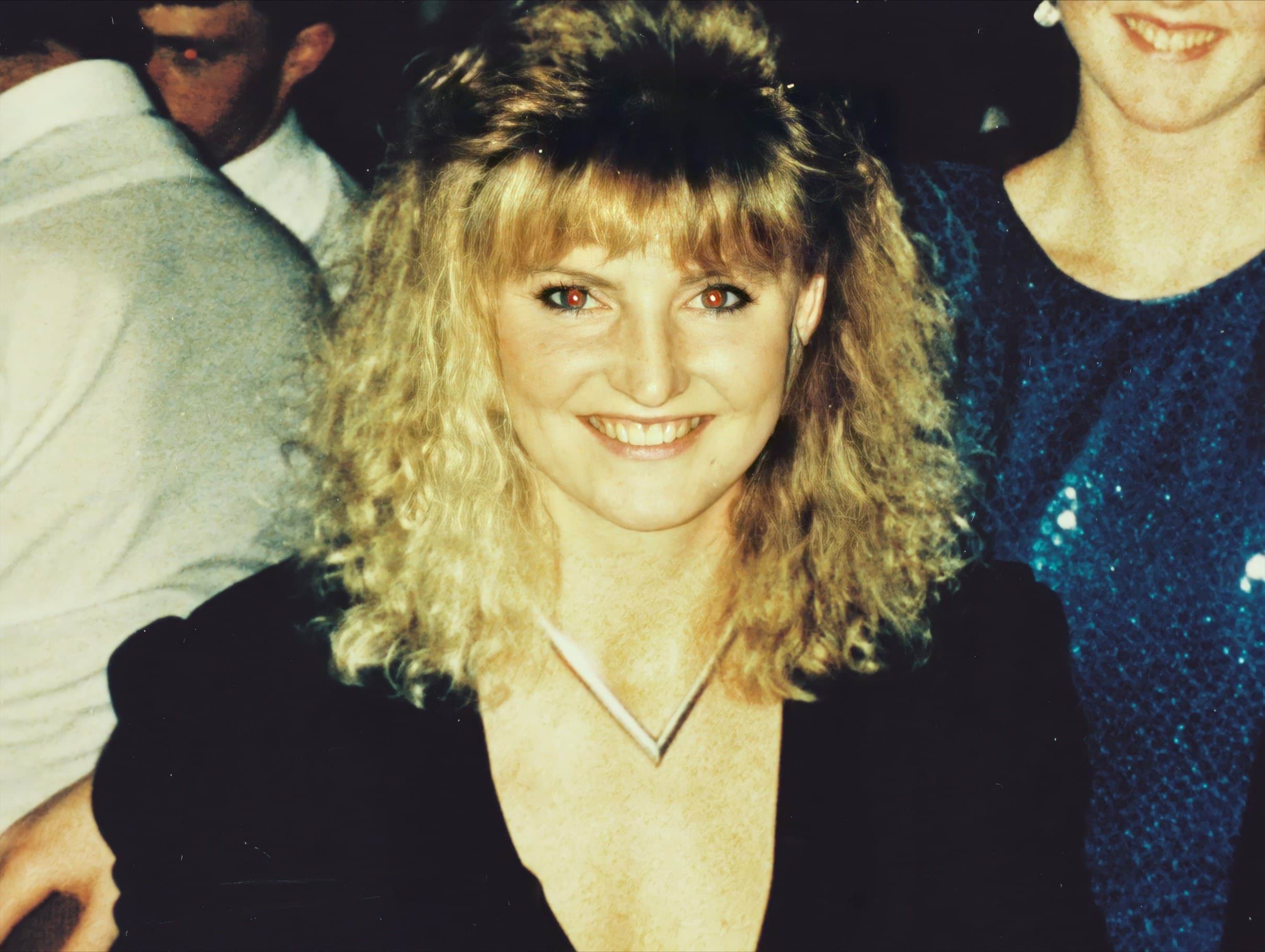 A 27-year-old white woman smiles directly at the camera. She has blonde curly hair, worn out at the sides and blow dried back at the front. She is wearing a black top and a distinctive silver v-shaped necklace. The photo looks to be from the early 1990's.