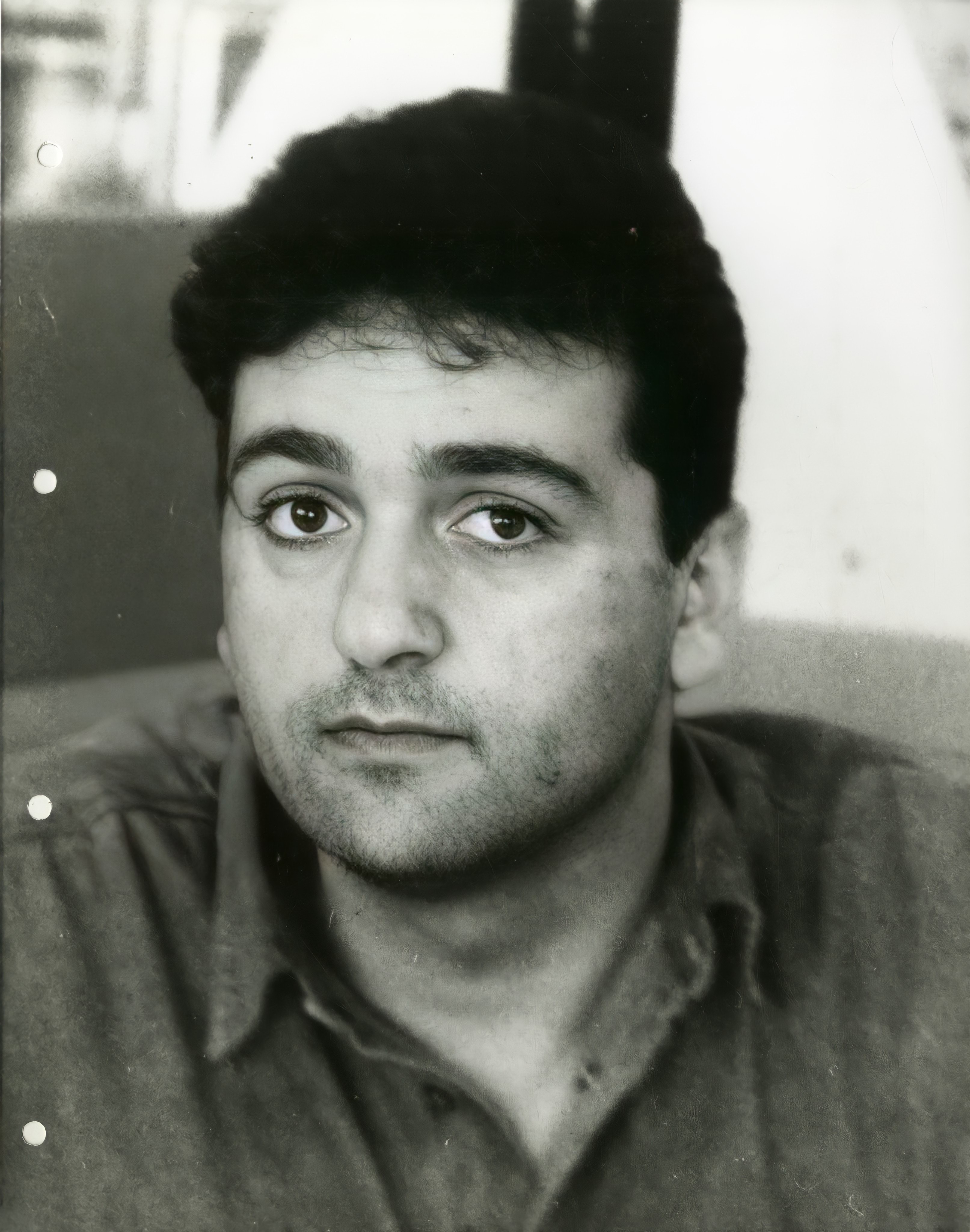 A black and white photo of 26-year-old white man looking directly at the camera. He has short dark curly hair, has slight stubble and dark eyes. The photo looks to be from the early 1990's.