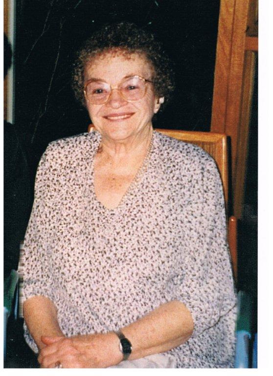 An older photo showing a older woman with very short and curly darker hair sits in a chair, smiling directly at the camera. Her hands are clasped in her lap, and she is wearing glasses, a watch and floral (or dotted) top. 