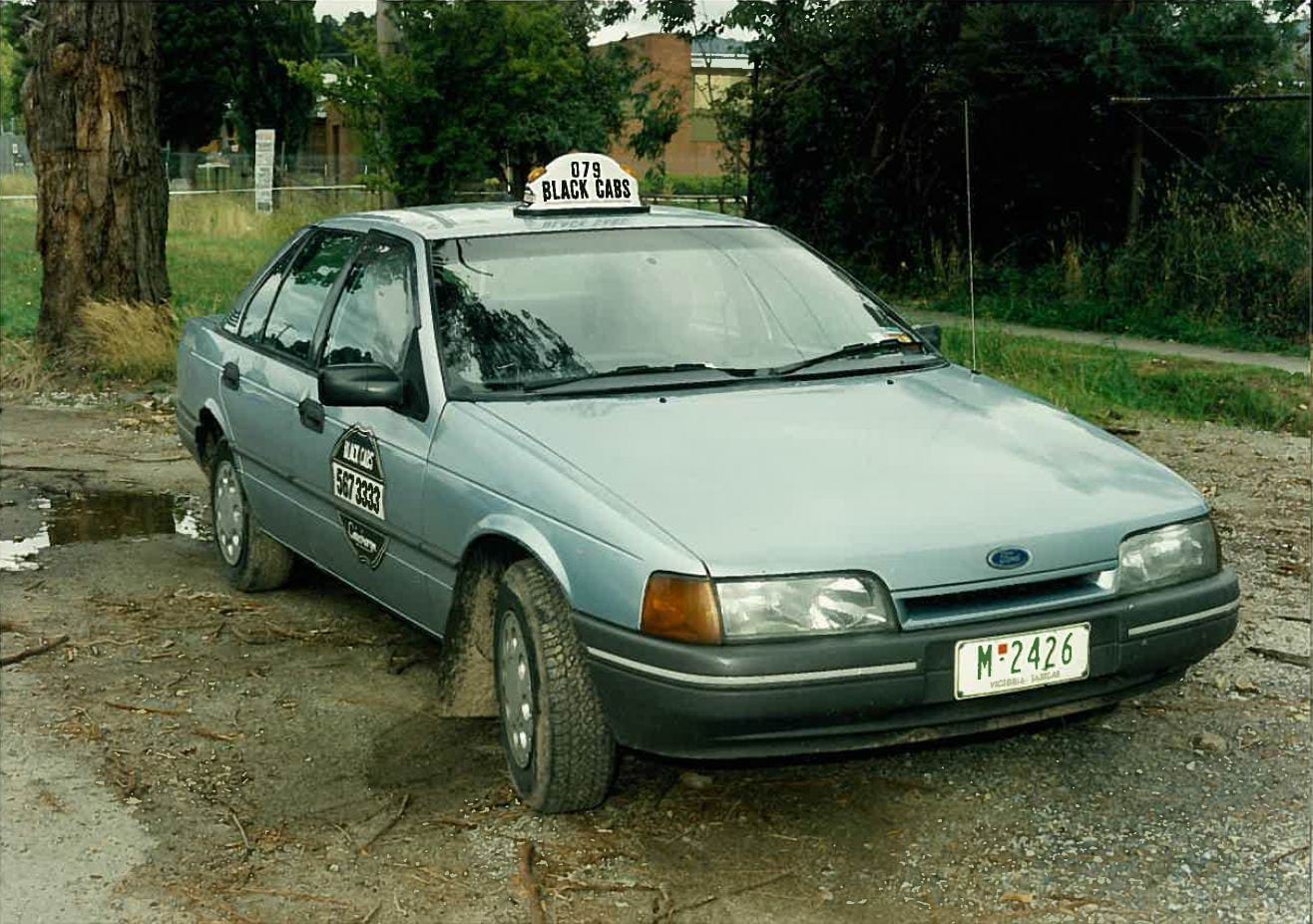 A blue grey early 90's taxi with the number plate M-2426 and a light on top with 079 Black Cabs. The cab is a Ford and is parked in a gravel carpark. 