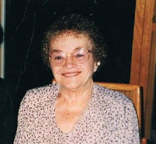 An older photo showing a older woman with very short and curly darker hair sits in a chair, smiling directly at the camera. Her hands are clasped in her lap, and she is wearing glasses, a watch and floral (or dotted) top. 