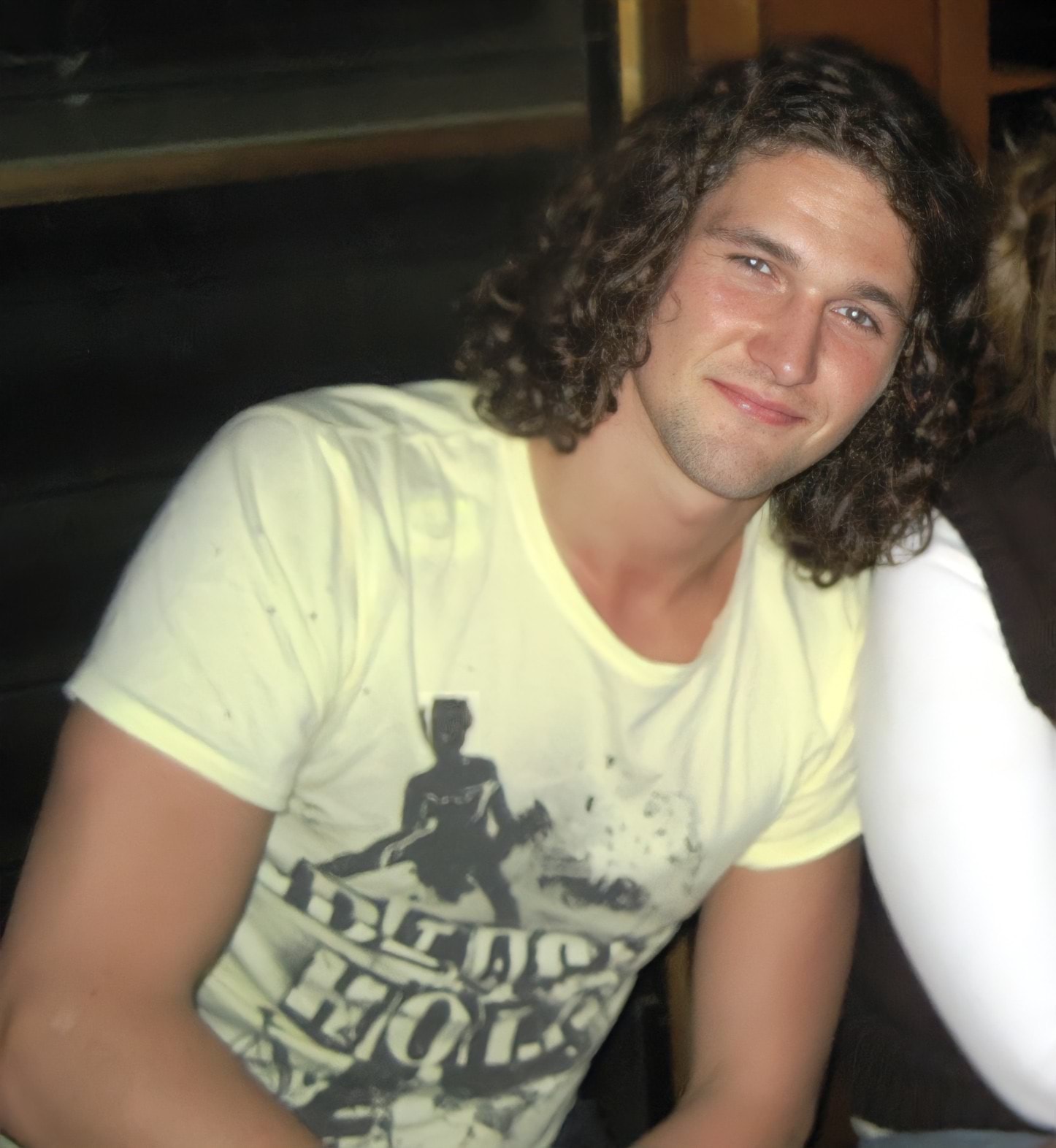 A 22-year old white man with mid-length brown curly hair is leaning to the side and smiling at the camera. He is wearing a pale yellow t-shirt and is leaning on the shoulder of someone sitting next to him. 