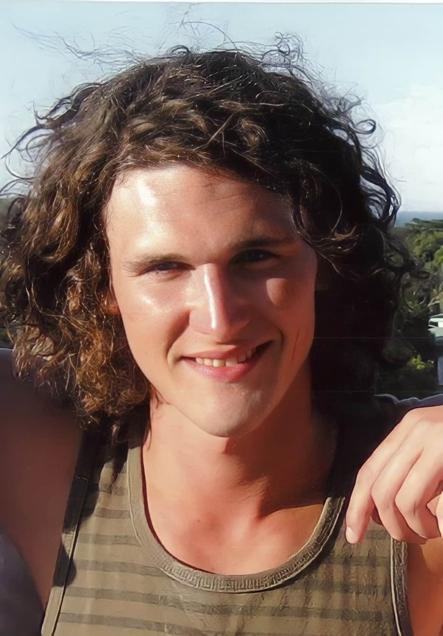 A 22-year old white man with mid-length brown curly hair smiles at the camera. He is wearing a brown striped tank top.