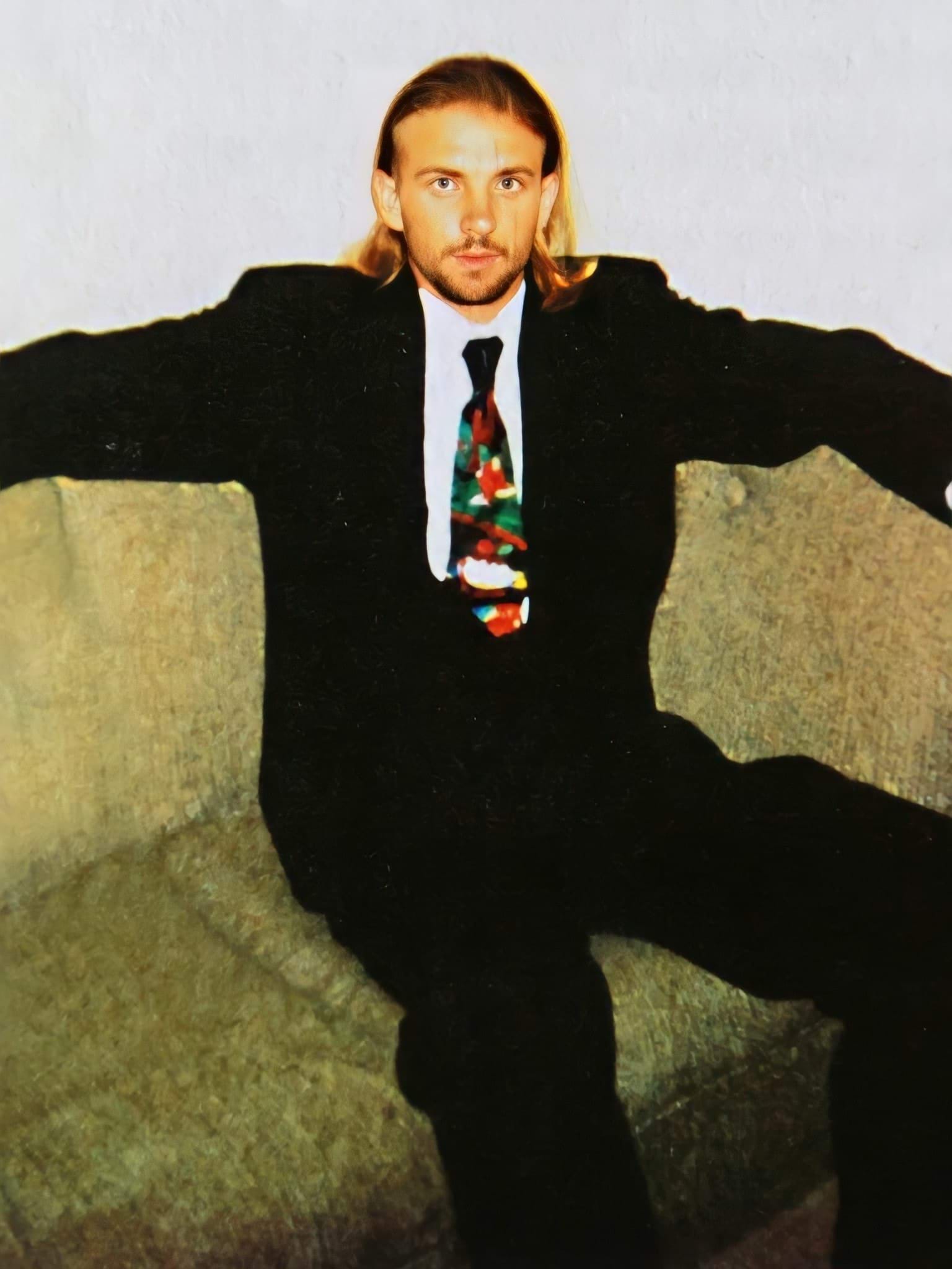 A 25-year-old white man smiles. He is wearing a wearing a black suit, white shirt and multi-coloured tie. He has shoulder length dark blonde hair and is looking directly at the camera. He is sitting on an olive coloured retro-style couch. The photo looks to be from the late 1990's.