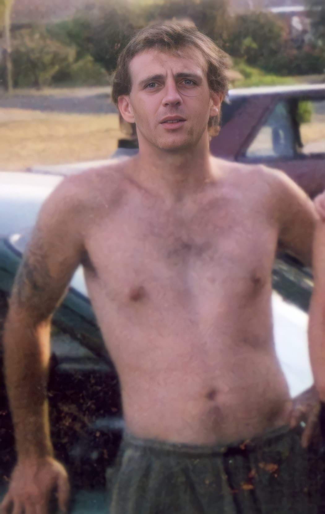 A 25-year-old white man is leaning back on the front of a car and looking at the camera. He has dark blonde/brown hair and is shirtless and wearing grey trackpants. There is a faint tattoo outline on his left bicep. The photo looks to be from the late 1990's or early 2000's.