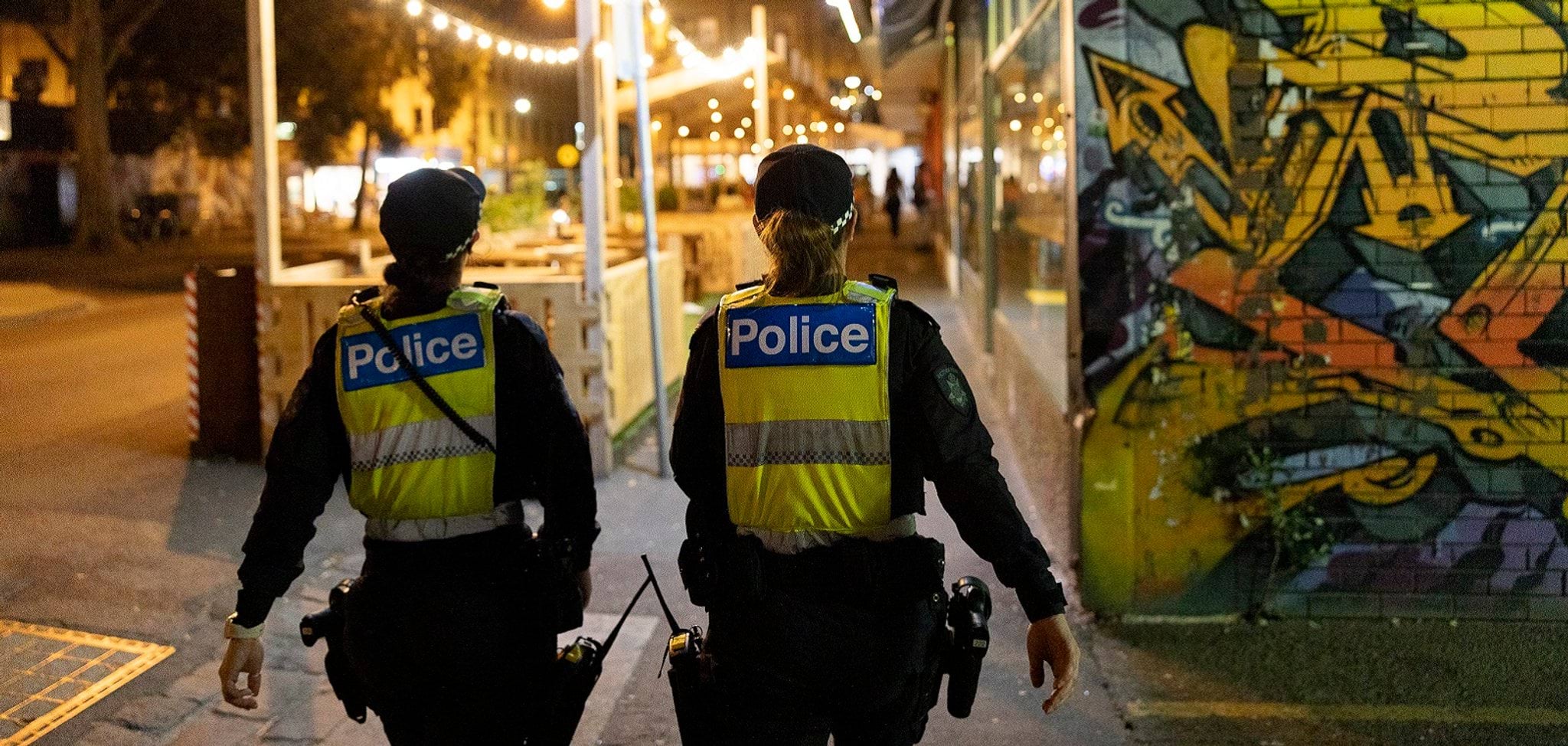 Two female police officers are walking down the street at night. They are facing away from us, and you can clearly read their high vis vests marked 'Police'. The street is brightly lit and you can see some graffiti on the wall next to them.