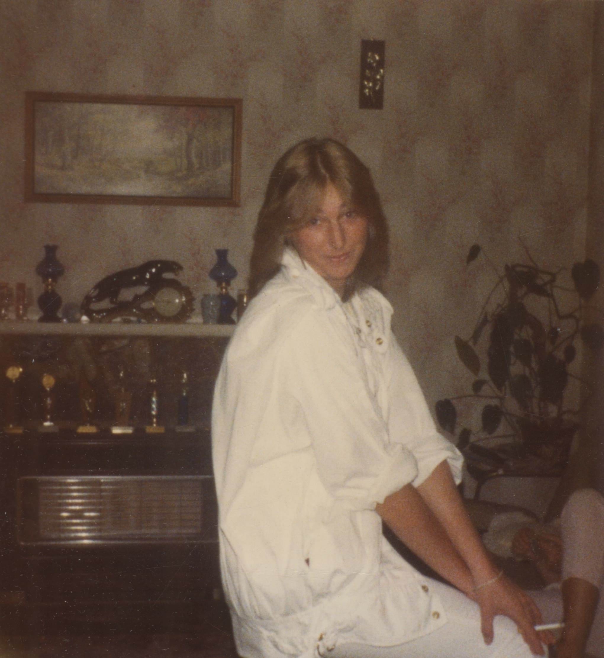 A 25 year old white woman with feathered dark blonde hair sits on a chair in a wall papered living room. She is looking down towards the camera, and is wearing a white parachute jacket and white pants. 