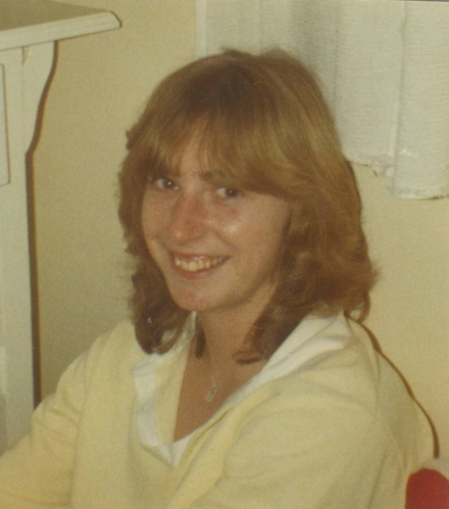 A 25 year old white woman with feathered dark blonde hair is sitting and smiling at the camera. She is wearing a pale yellow polo shirt with a white collar.