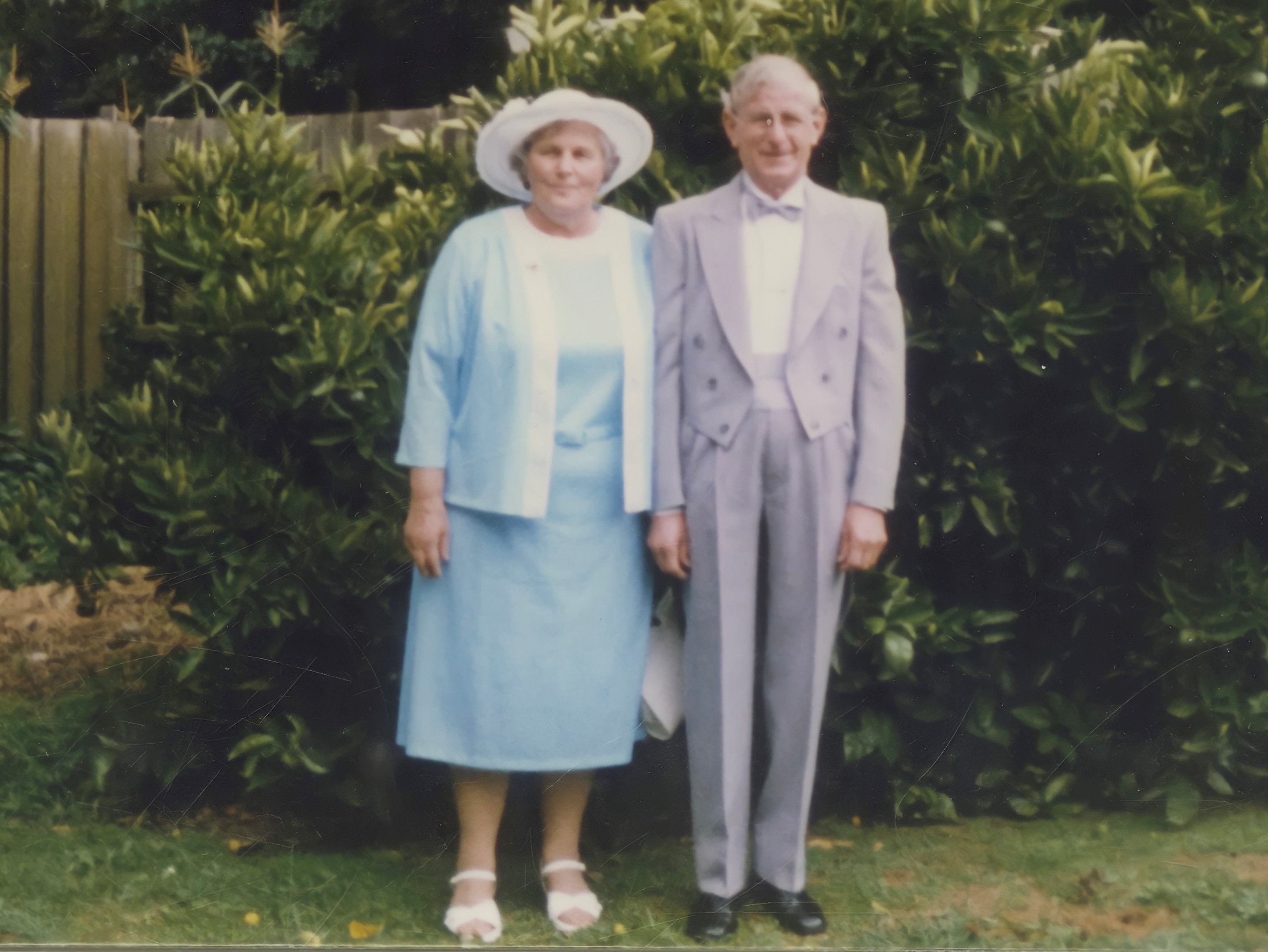 A 64-year-old white man is standing next to his wife. They are both dressed formally. His has silver white hair and is wearing a pale purple tuxedo. She is wearing a pale blue dress. The are standing in a garden and looking at the camera.  