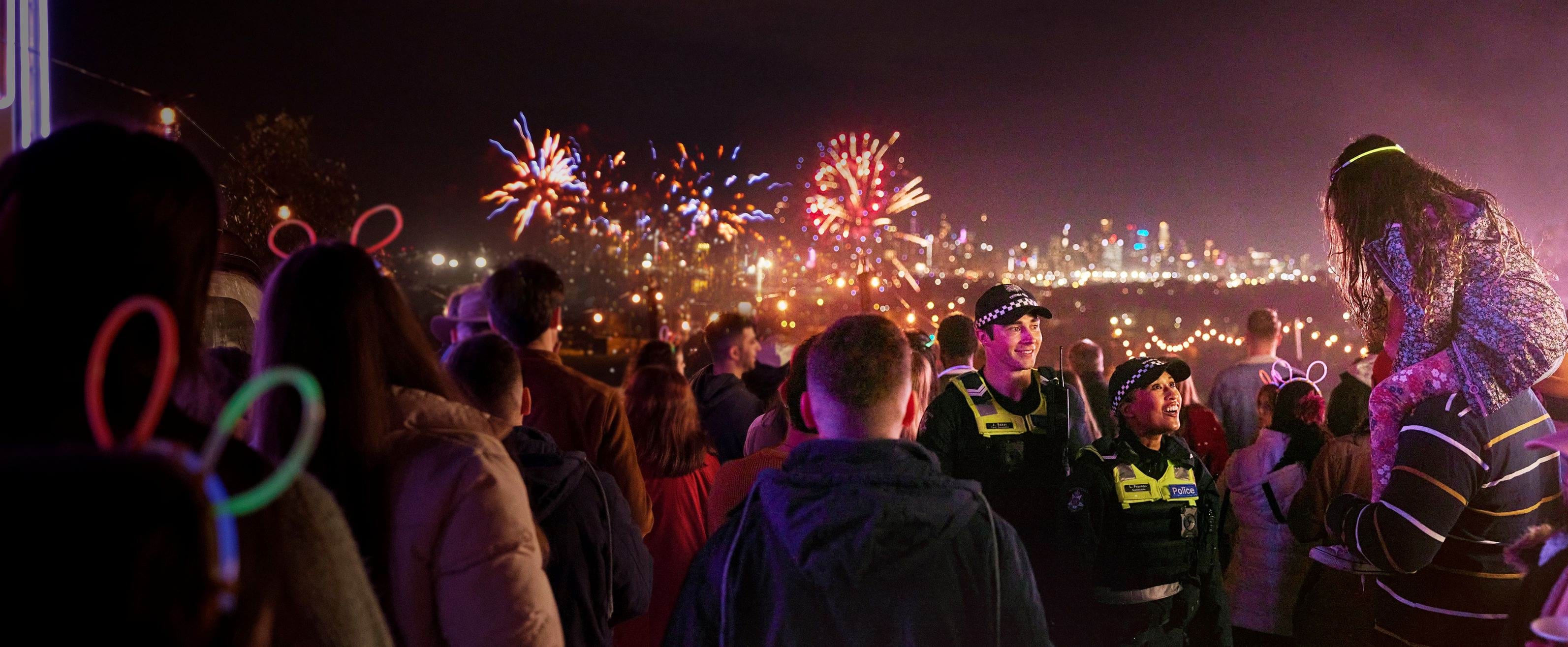 Two officers in a crowd at a night carnival. The crowd is watching fireworks in the distance, but the two uniformed officers are looking towards the camera. 