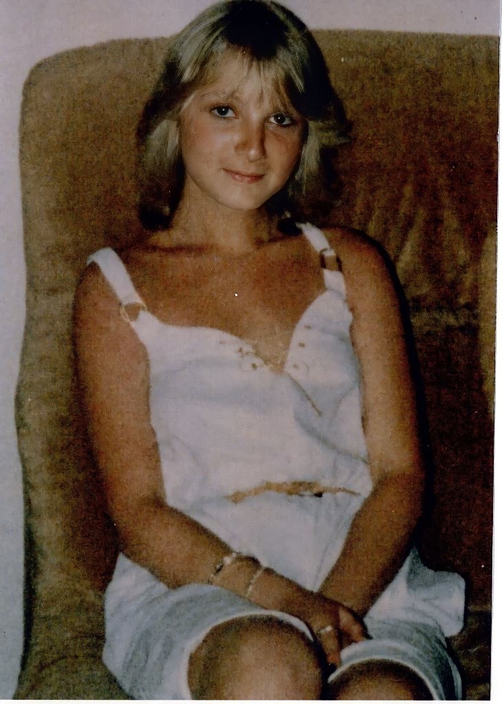 A 17-year-old Caucasian woman is smiling slightly and looking directly at the camera. She has short, shoulder-length dark blonde hair with a fringe that sits slightly below her eyebrows. She is wearing a white, sleeveless dress with a lace up detail at the front, silver bangles, a silver ring and a brown belt. She is sitting on a brown couch. The photo looks to be from the mid-1980’s.