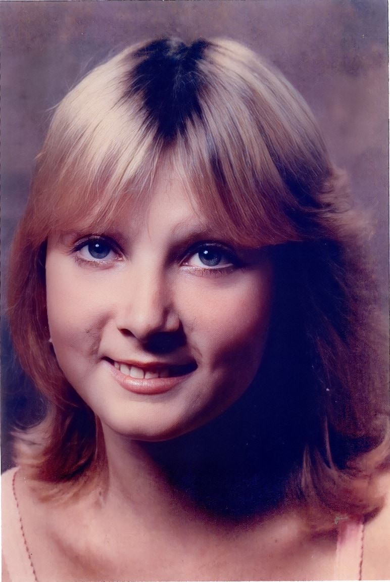 A 17-year-old Caucasian woman is smiling and looking directly at the camera. She has dark blonde hair with brown regrowth showing at the top of her head. She has blue eyes. The photo looks to be from the mid-1980’s.