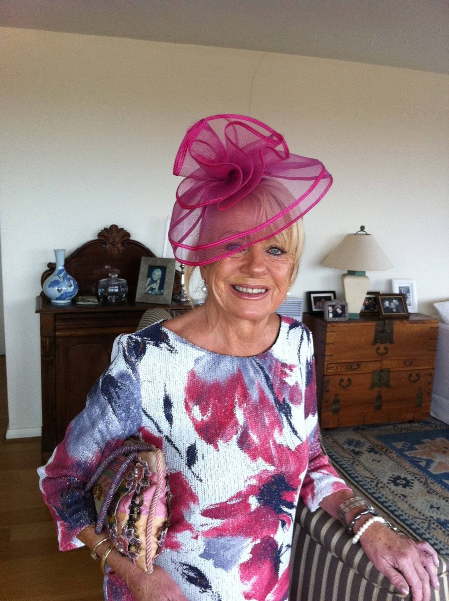 A 69-year-old Caucasian woman with blonde hair is looking directly at the camera and smiling with teeth. She is wearing a pink fascinator which obscures her right eye, and a white 3/4 quarter length sleeved white dress with pink and purple flowers on it. She has blue eyes and is wearing black eyeliner.