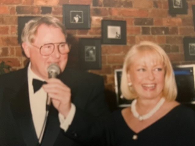 A 69-year-old Caucasian woman is looking away from the camera and smiling. She has blonde, shoulder-length hair and blue eyes. She is wearing a white necklace and a black top. She is standing next to a Caucasian man of approximately the same age wearing a tux, bowtie and glasses. He is holding a microphone and looking away from the camera.