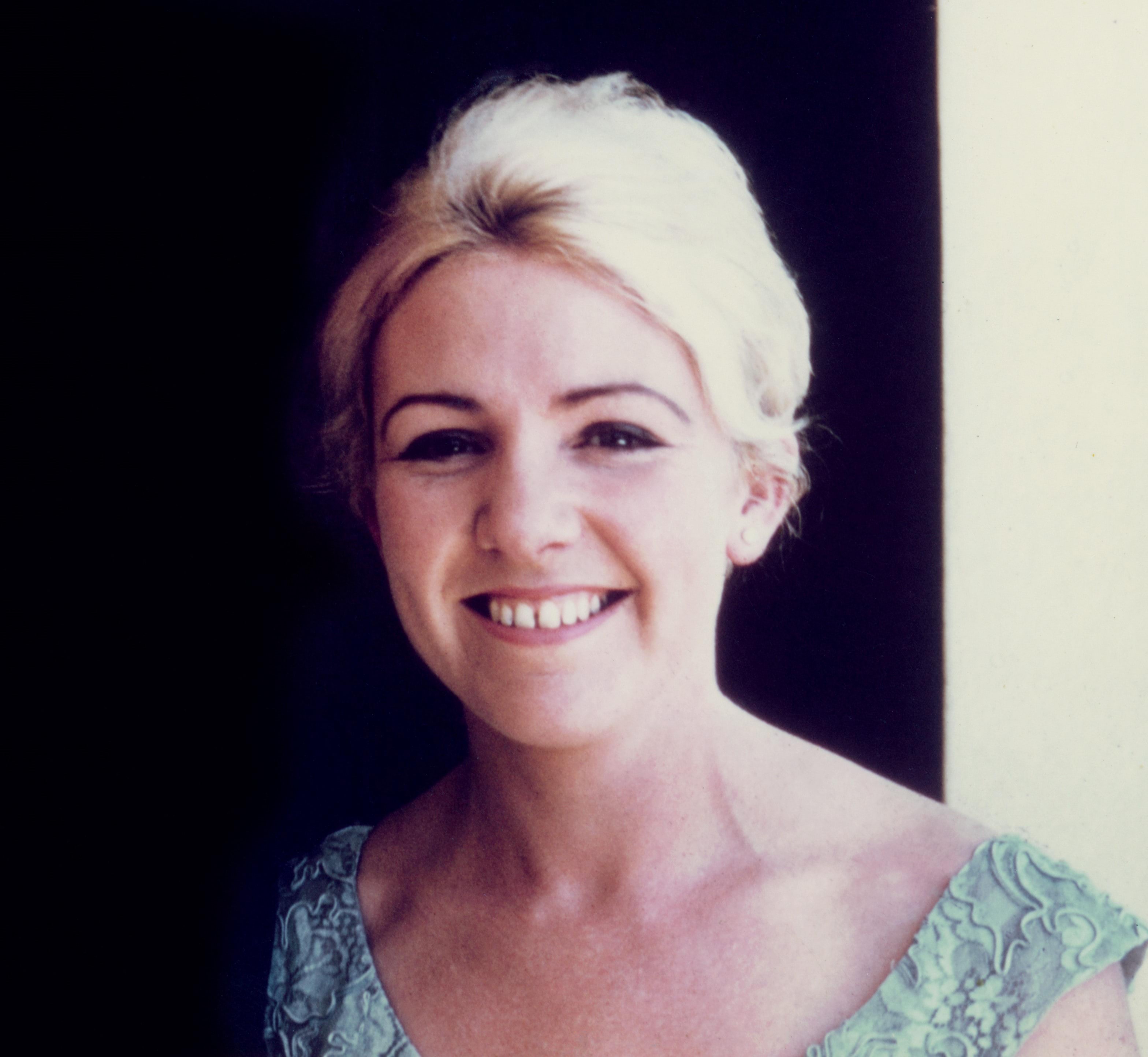 A 41-year-old Caucasian woman is smiling widely and looking directly at the camera. She has dyed, white blonde hair tied back away from her face. She has blue eyes and thin brown eyebrows. She is wearing a blue top with lace, floral detail. The photo appears to be from the mid 1970's.