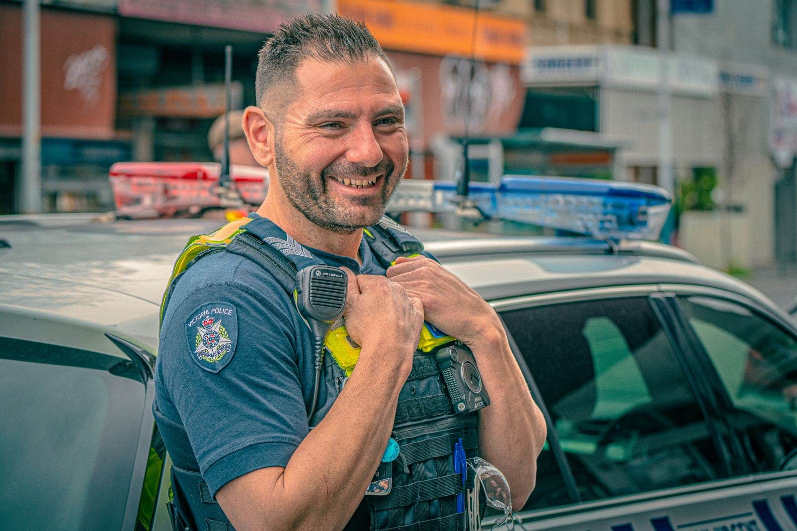 A Victoria Police police officer stands in front of a Victoria Police car on a Melbourne street. He is smiling widely and holding his arms to his chest.