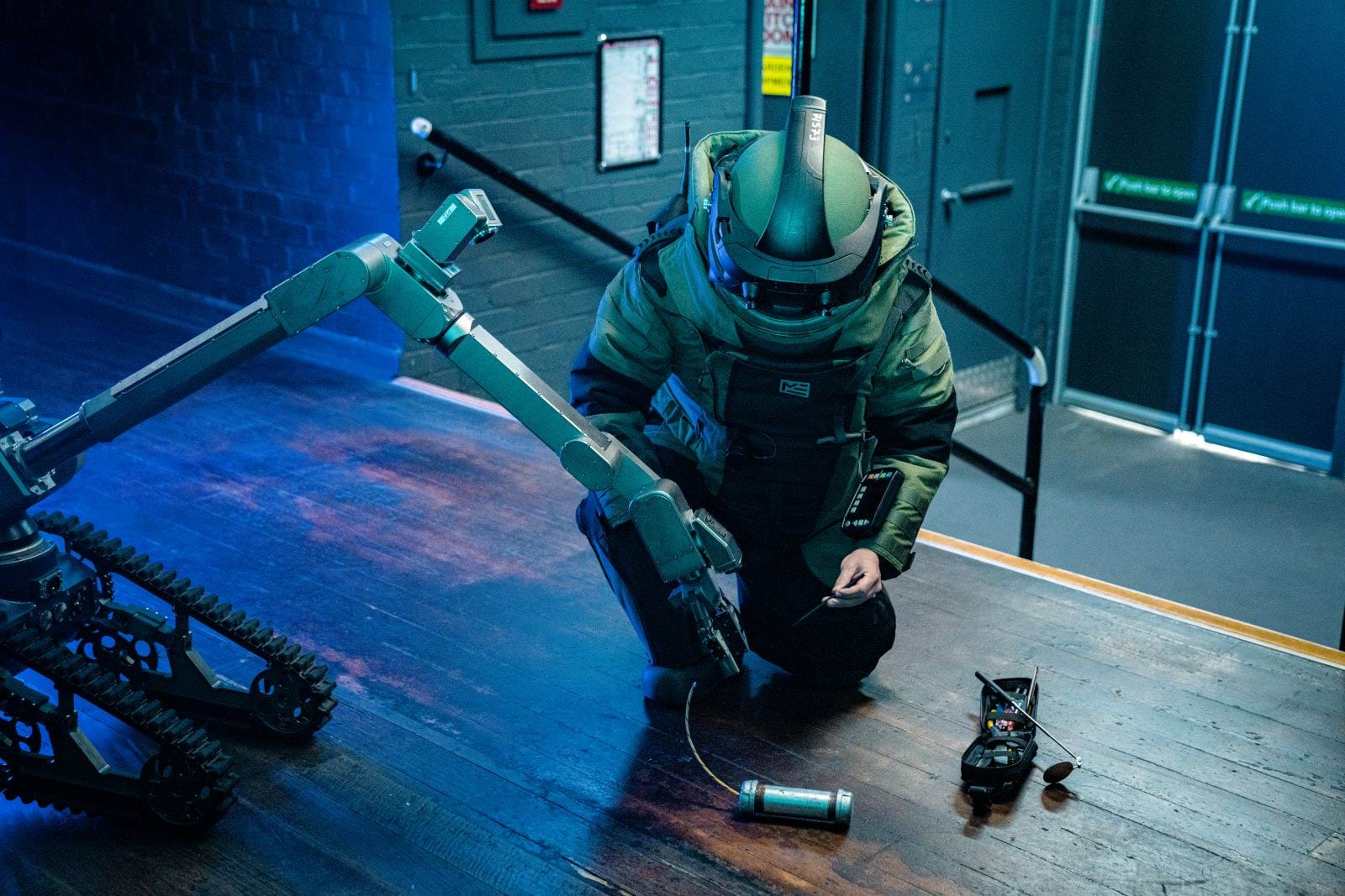 Bomb squad officer in personal protective gear, dismantling a bomb with the assistance of a bomb defusal robot, in a test environment. 