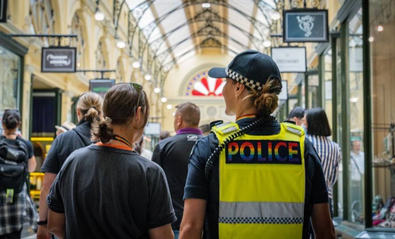 Valuing diversity at Victoria Police From the back - officer with Victoria Police pride logo high vis vest on and a member of public walking inside a shopping centre.