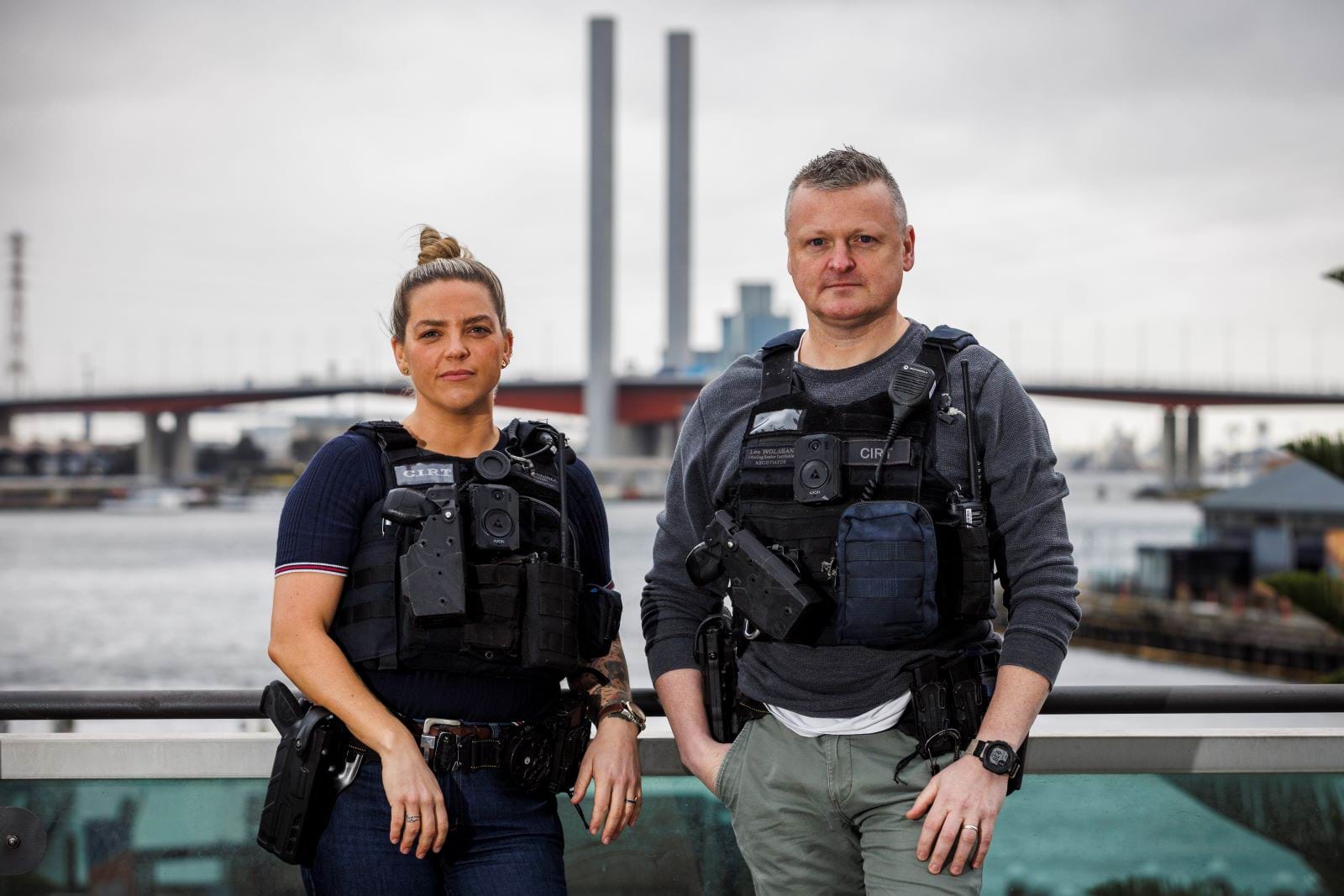 A female and male negotiator in Victoria Police’s Critical Incident Response Team (CIRT), dressed in civillian clothes but armned, with CIRT tactical vests over the top of their clothes. The negotiators are unsmiling and facing the camera, and standing with a the Docklands waterfront and a large traffic bridge in the distance behind them. 