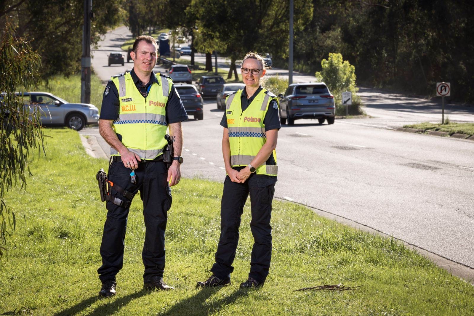 Two officers from the  Major Collision Investigation Unit stand on the grass next to a two lane road. They are looking directly at the camera and wearing bright reflective police vests. There are cars in the background on the road behind them.