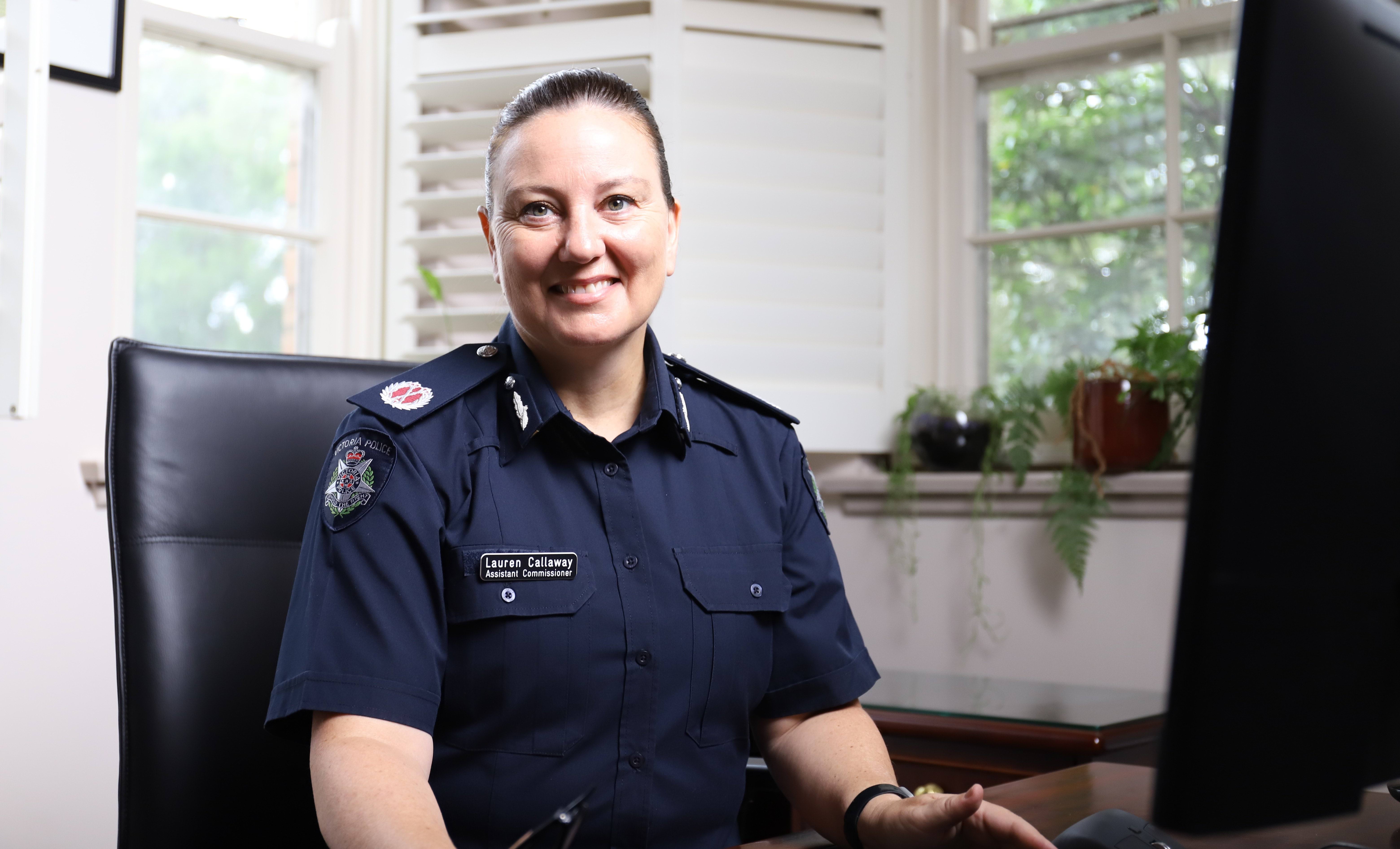 Assistant Commissioner Lauren Callaway is wearing a navy police uniform. She is sitting at a desk, smiling at the camera. 