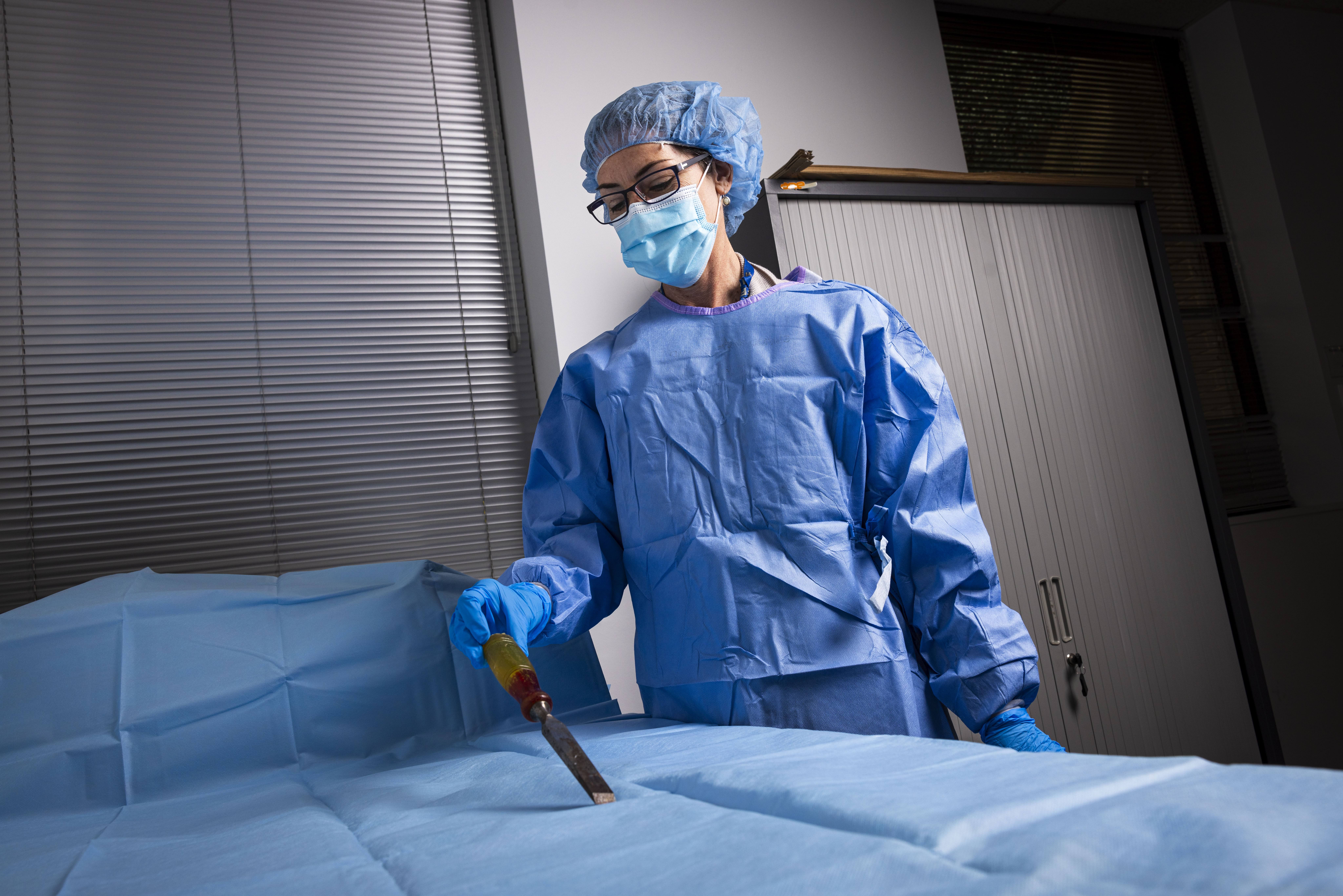 Forensic Scientist Kate Outteridge is wearing a protective blue cap, mask, gloves and gown. She is examining a piece of evidence on a blue sterile sheet.