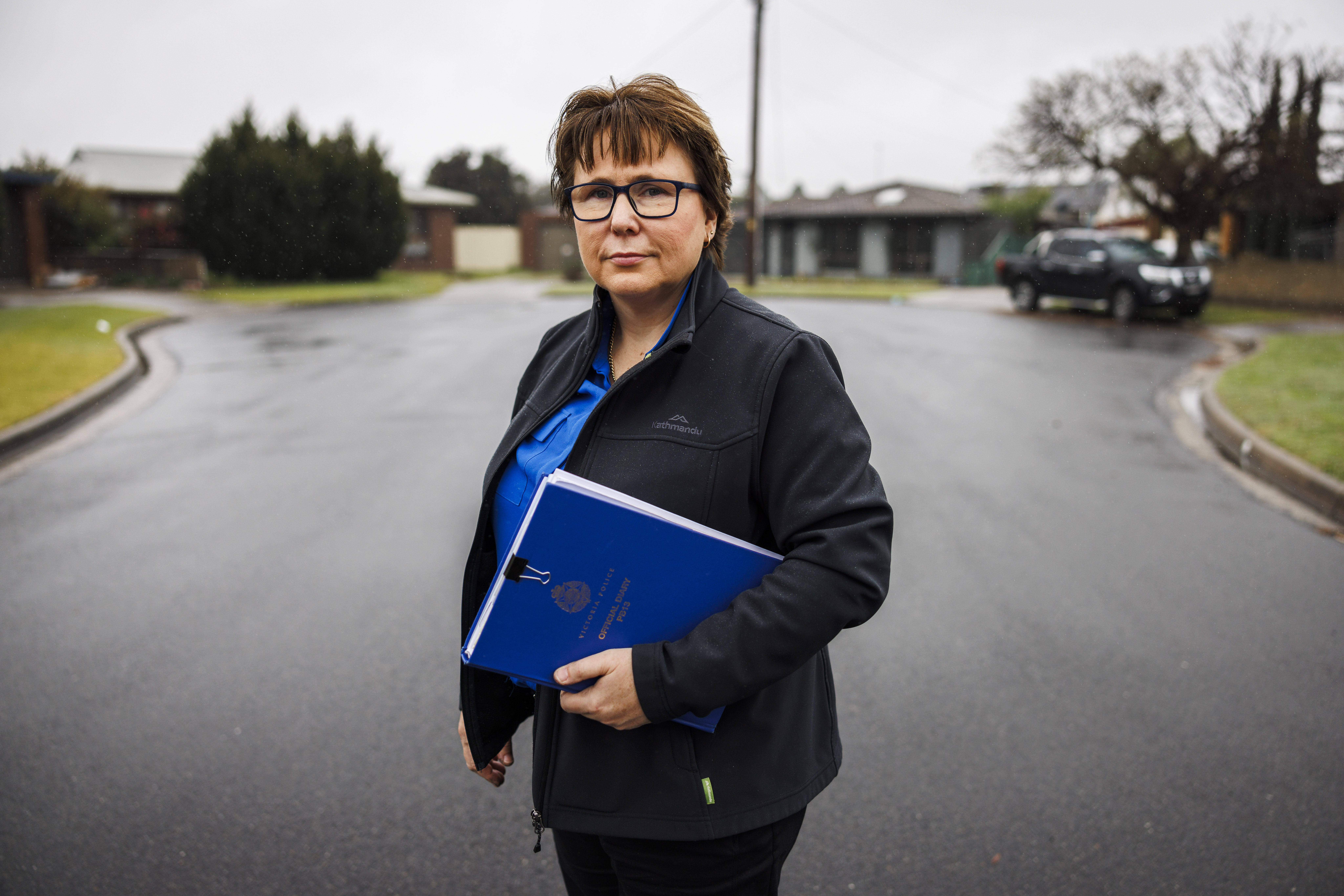 Detective Leading Senior Constable Tracy Jarrott is standing on the road, looking at the camera. She is holding a blue Victoria Police Official Diary.