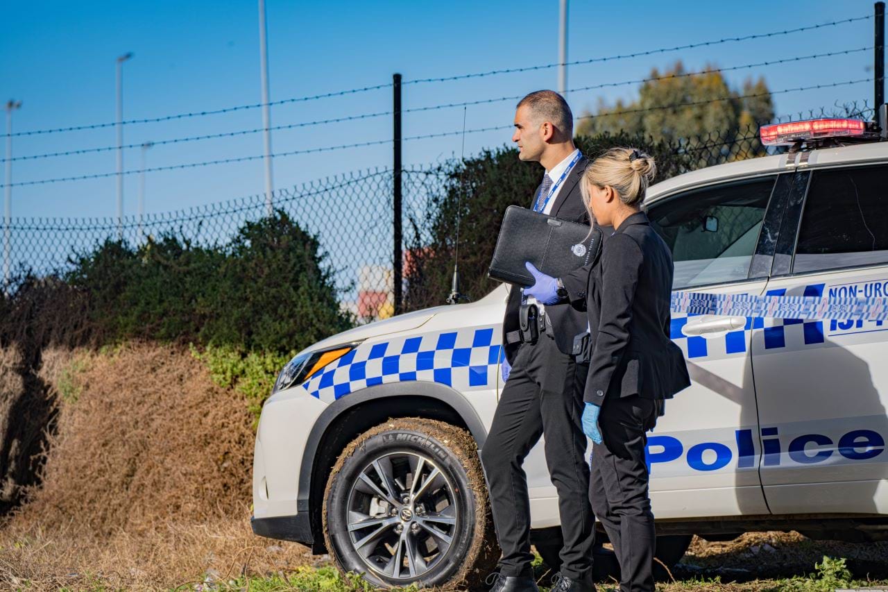 Two detectives in suits stand next to a police vehicle while looking into the distance. They are both wearing gloves, and one is holding an official looking folder. 