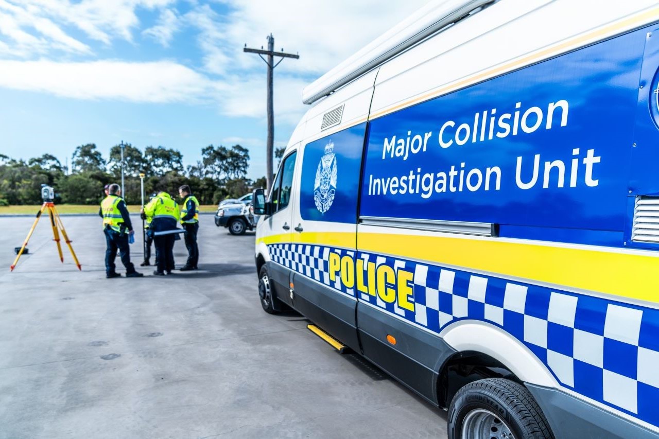 A group of Major Collision Unit officers stand in a car park, having a discussion with some equipment around them. A branded MCIU police van is parked facing them. 