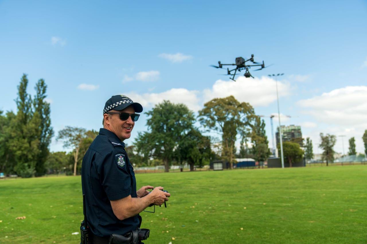 A uniformed RPAS officer operates a drone in a green field, and you can see the drone flying in the background. The officer has turned to face the camera and is wearing sunglasses and smiling. 