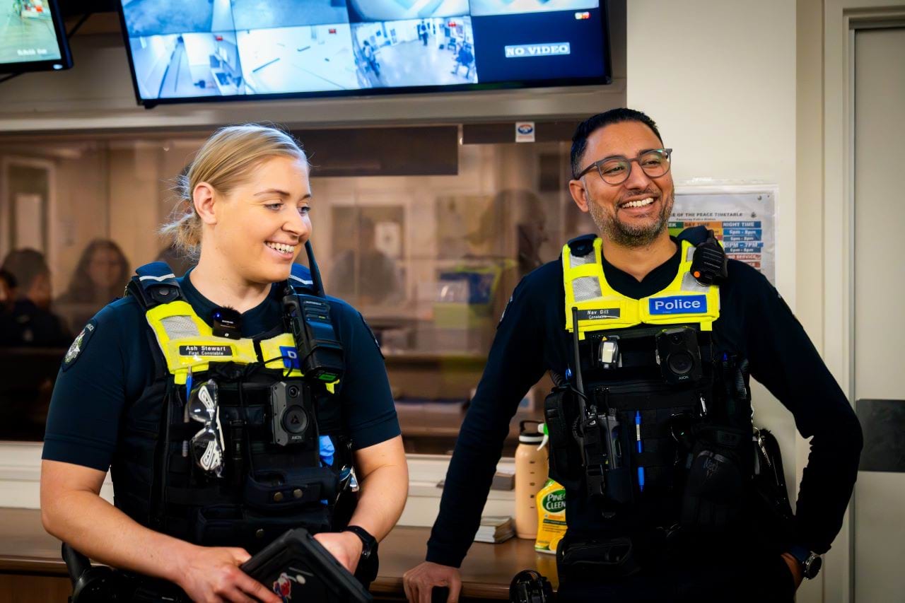 Two officers, in full uniform and gear, are smiling while in a local police station. There are some digital screens behind them, and one is leaning with a hand on a desk behind them. If the background, another officer at the front counter talks to a community member.  