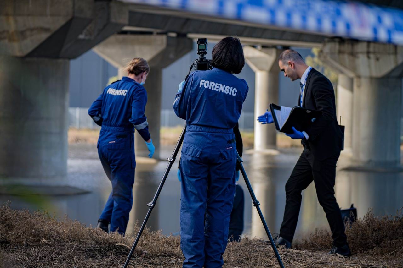 A forensic services officer in blue coveralls uses equipment to survey a crime scene. There is another forensics officer and a detective on either side investigating, and a body of water in the background. They appear to be under a bridge. 