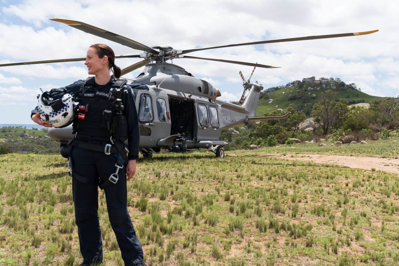 An airwing officer, in flight gear, stands in a field in front of a police helicopter. They are holding a police branded flight helmet, looking to the side and smiling. 