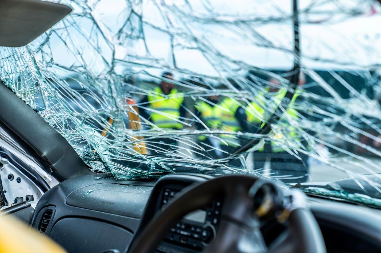 A group of Major Collision Unit officers can be seen distorted through a smashed car window.  They are wearing high visibility gear and appear to be discussing the damaged car.