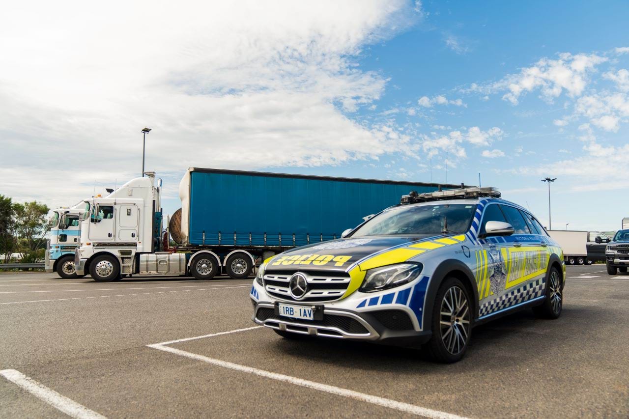 A Heavy Vehicle Unit branded police station wagon is parked in a car park. In the background two large trucks can be seen parked next to each other.