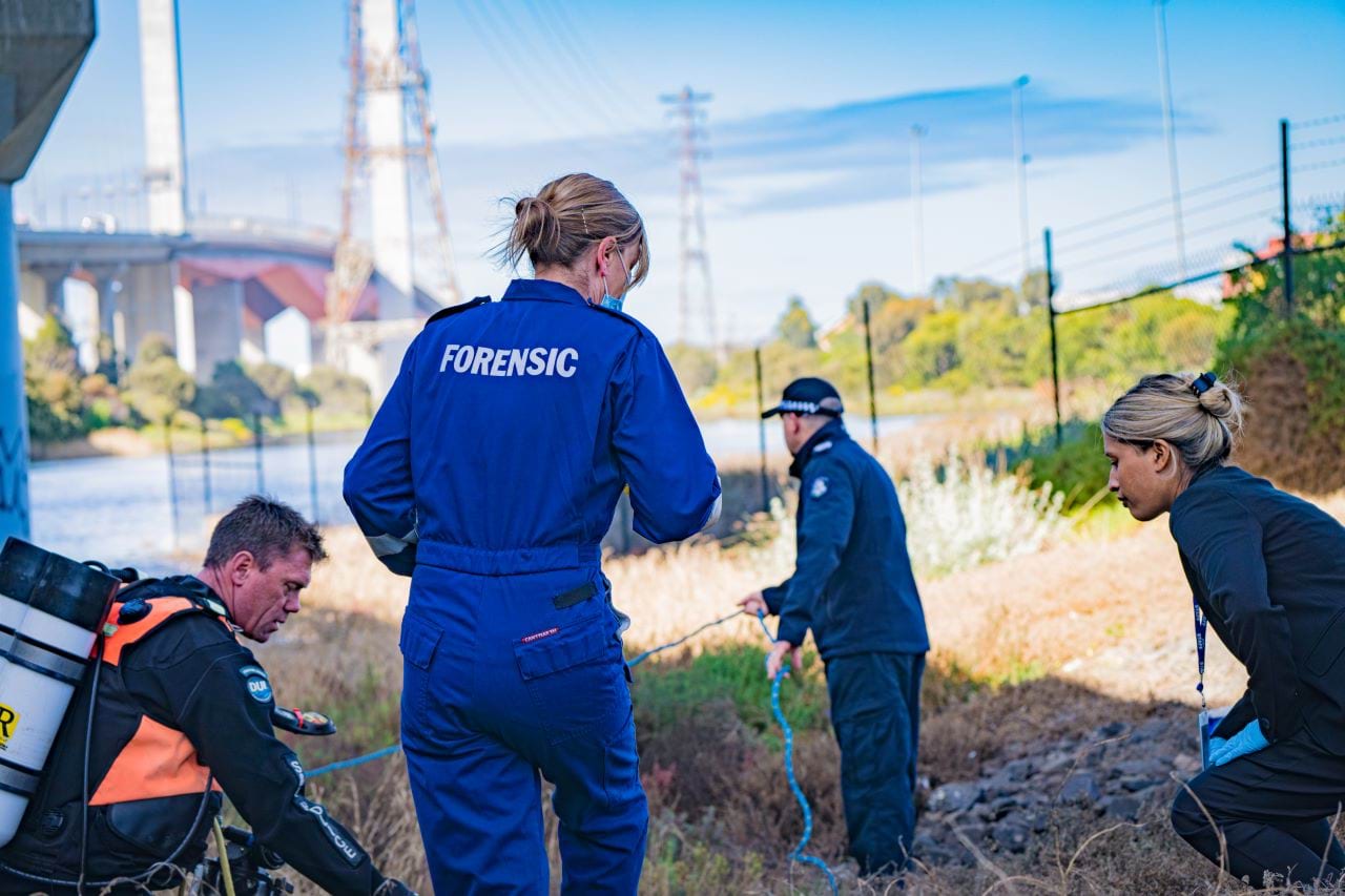 A Forensic team is investigating an outdoor crime scene. They appear to be under a bridge, with a body of water just behind them and the Bolte Bridge in the distance. A forensic officer is surveying the ground, with a detective and a diver crouched either side. A uniformed police officer appears to be pulling a rope in the background. 