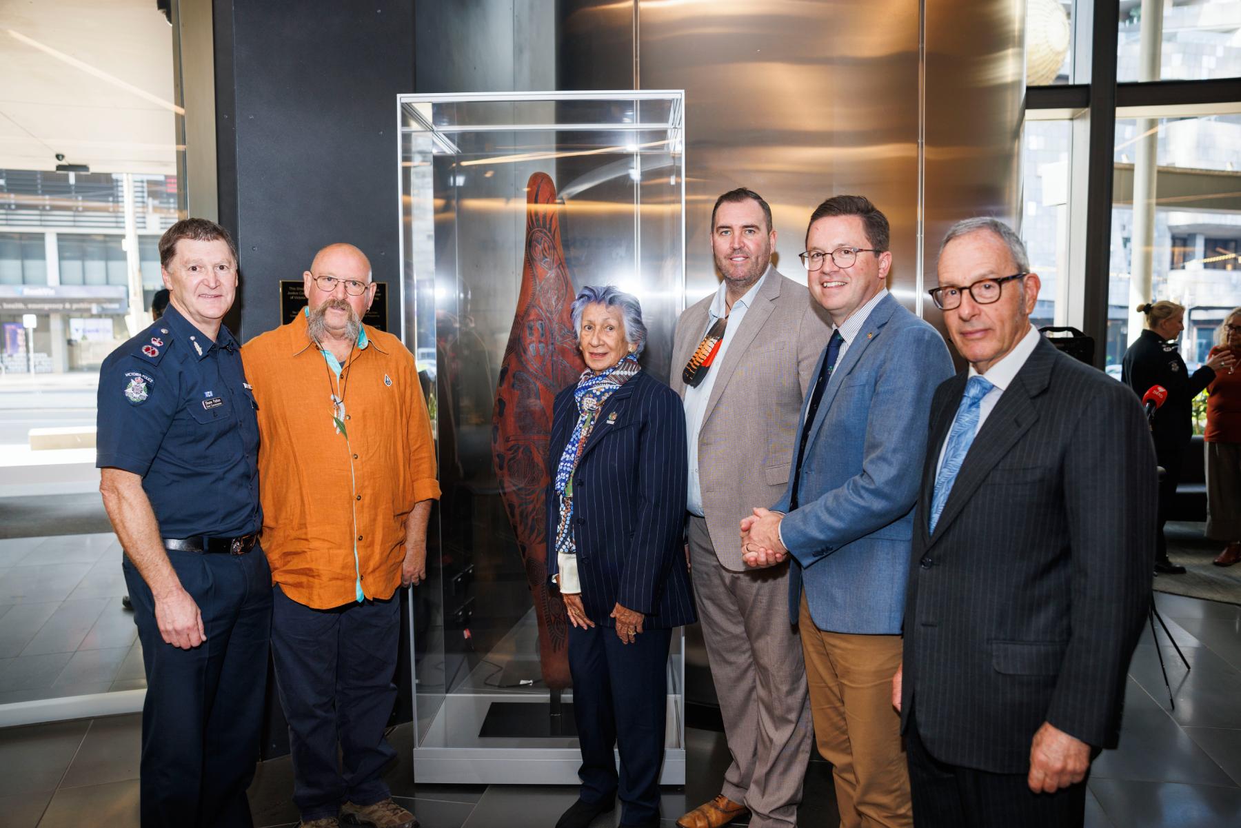 Left to right: Chief Commissioner of Police, Shane Patton APM, Uncle Mick Harding, Yoorrook Justice Commission Chair Professor Eleanor Bourke AM, Yoorrook Justice Commissioner Travis Lovett, Hon Anthony Carbines MP, Minister for Police, Yoorrook Justice Commissioner The Hon. Anthony North KC in the foyer of the Victoria Police Centre. The Shield is made of wood with intricate Aboriginal carvings and is displayed in a glass cabinet central to the image.