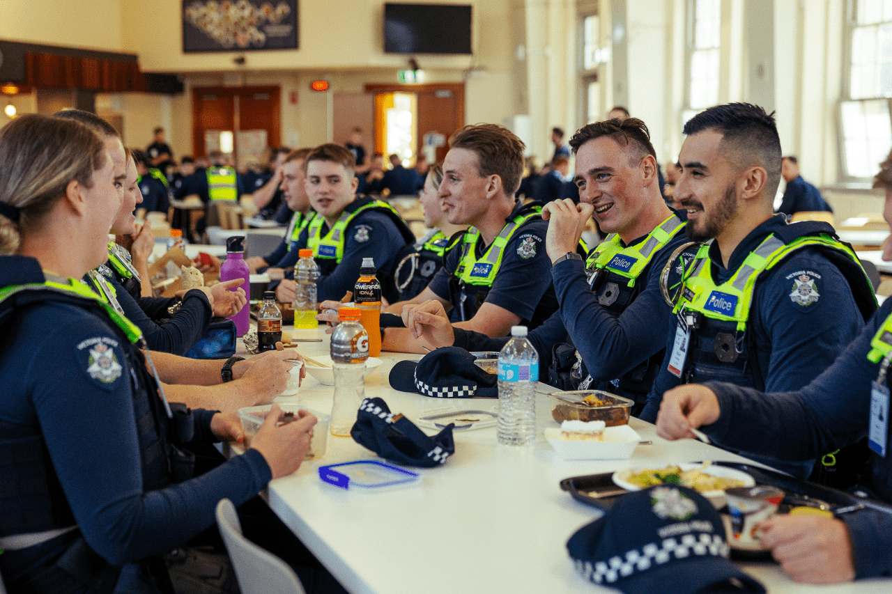 A group of Victoria Police recruits are having lunch together in the cafeteria at the Victoria Police Academy. There is a big group of them sharing a table in uniform, chatting and smiling. 