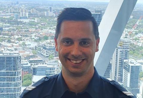 Senior Constable Joshua James, a proud Noongar man, is shown smiling and looking at the camera. He is wearing his Victoria Police uniform, and a view of the Melbourne Central Business District is visible out the window behind him.